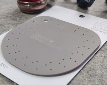 Puede incluir: Una herramienta de dobladillo de esquina de silicona gris con un ángulo de 230 grados. La herramienta tiene una cuadrícula de pequeños agujeros para presionar la tela.