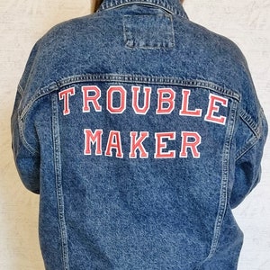 Puede incluir: Una chaqueta vaquera azul con las palabras "TROUBLE MAKER" bordadas en la espalda en rojo y blanco.