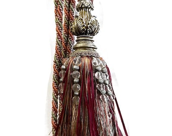 May include: An ornate curtain tieback with a decorative tassel. The tassel features a mix of burgundy, gold, and silver threads, accented with clear, faceted beads. The braided cord is in shades of brown, green, and orange.