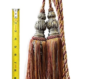 May include: Two tassel tiebacks with gold, red, and cream colored cords. The tassels have silver metal tops with a decorative design. The cords are twisted together.