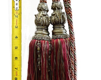 May include: Two decorative curtain tassels with ornate, bronze-colored tops and burgundy fringe. The tassels are accented with gold and clear beads. The twisted cords are brown, green, and orange. The tassels are approximately 9 inches long.