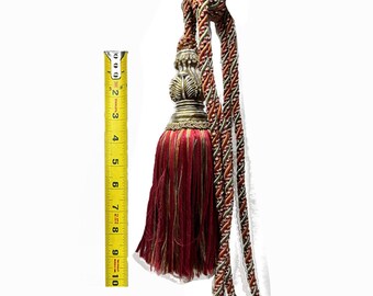 May include: A red and gold tassel with a decorative metal top. The tassel is approximately 5 inches long and is attached to a cord.