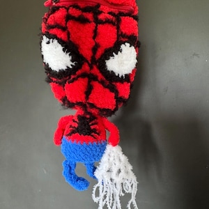 May include: A red and blue tufted pouch shaped like a character head with a zip closure at the top. The design features a red face with black web detailing, large white eyes, a black spider emblem on the chest, and a white web-like fringe.
