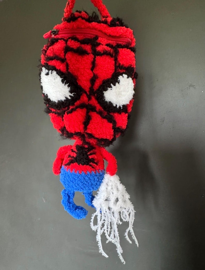 May include: A red and blue tufted pouch shaped like a character head with a zip closure at the top. The design features a red face with black web detailing, large white eyes, a black spider emblem on the chest, and a white web-like fringe.