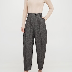 May include: A woman is wearing a light beige turtleneck sweater and dark grey high-waisted trousers with a subtle herringbone pattern. She is wearing brown leather loafers with a buckle.