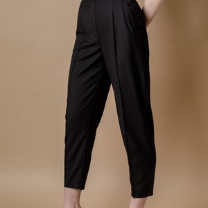 May include: Black high-waisted pants with pleats and a button closure. The pants are cropped and have a straight leg. The model is wearing the pants with nude heels.