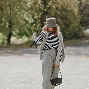 May include: A woman wearing a gray bucket hat, a striped shirt, a gray cardigan, gray pants, and black shoes. She is carrying a black shoulder bag.