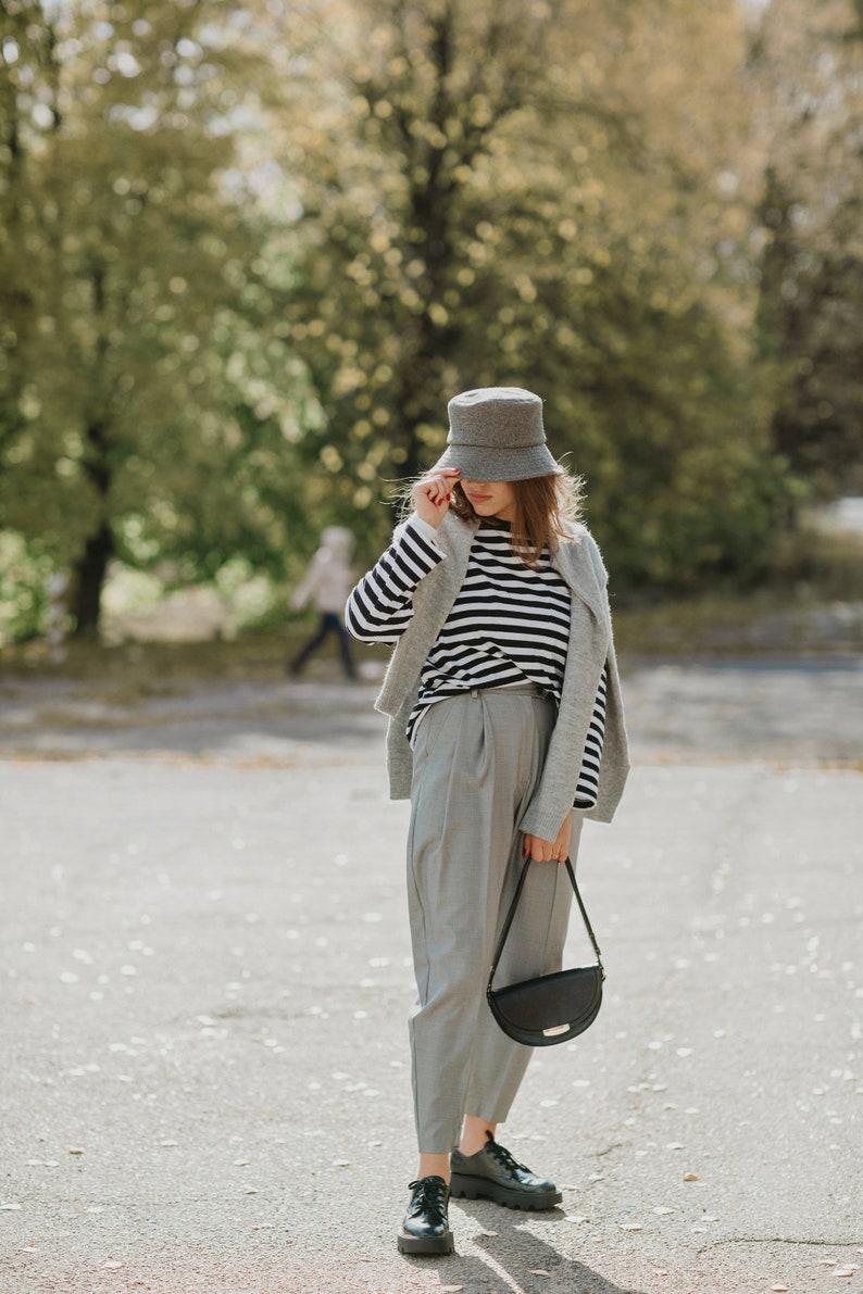 May include: A woman wearing a gray bucket hat, a striped shirt, a gray cardigan, gray pants, and black shoes. She is carrying a black shoulder bag.