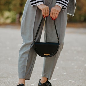 May include: A woman wearing gray pants, a striped shirt, and black leather shoes is holding a black half-moon shaped handbag with a gold clasp.