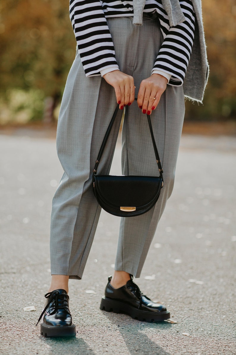 May include: A woman wearing gray pants, a striped shirt, and black leather shoes is holding a black half-moon shaped handbag with a gold clasp.