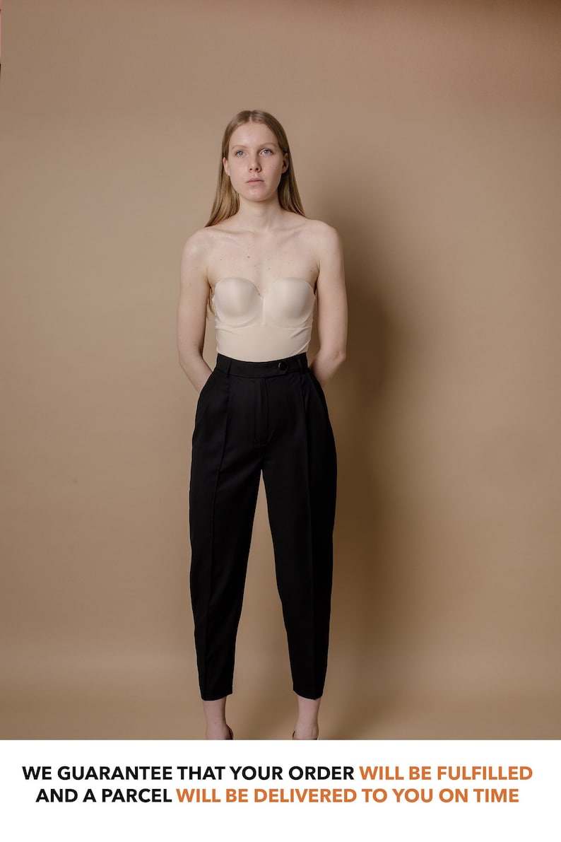 May include: A woman wearing a strapless beige top and black pants. The background is a light brown color.