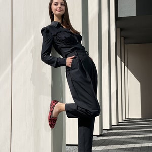 May include: A woman wearing a black ruffled blouse and black pants with a red and black leopard print flat shoe.