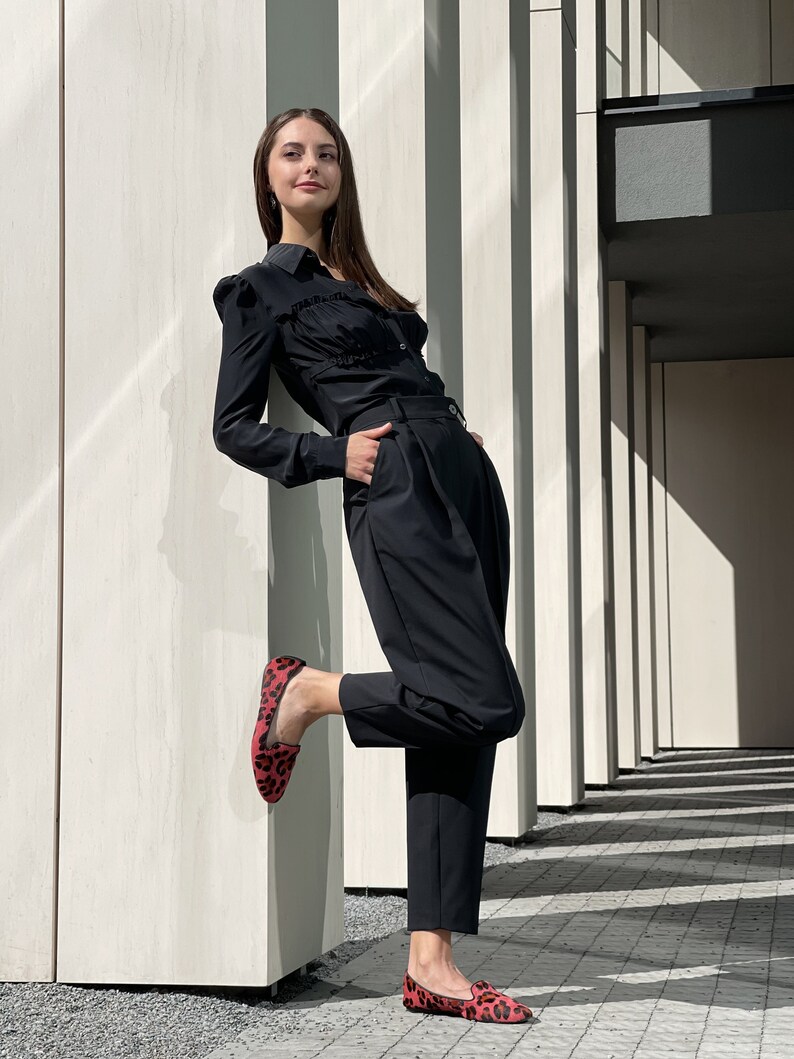 May include: A woman wearing a black ruffled blouse and black pants with a red and black leopard print flat shoe.