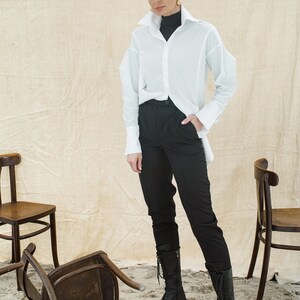 May include: A woman wearing a white oversized button-down shirt, black pants, and black lace-up boots. She is standing in front of a beige background with three wooden chairs.