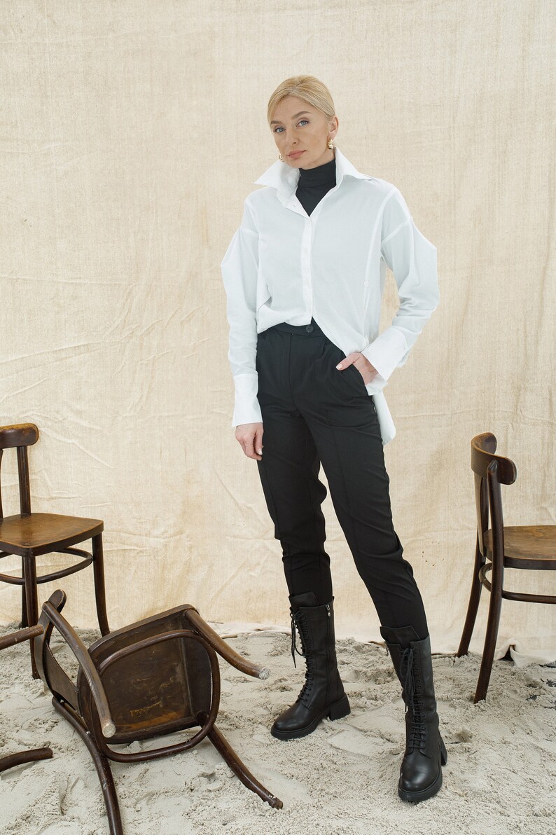 May include: A woman wearing a white oversized button-down shirt, black pants, and black lace-up boots. She is standing in front of a beige background with three wooden chairs.