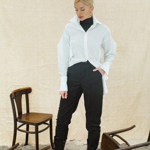 May include: A woman wearing a white button-down shirt over a black turtleneck, black pants, and black lace-up boots. She is standing in a room with several wooden chairs overturned on the floor.
