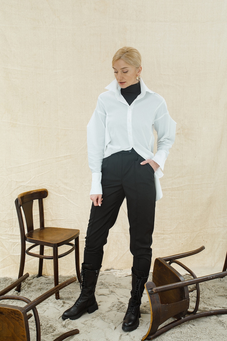 May include: A woman wearing a white button-down shirt over a black turtleneck, black pants, and black lace-up boots. She is standing in a room with several wooden chairs overturned on the floor.