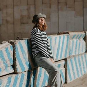 May include: A woman wearing a gray hat, black and white striped long-sleeve shirt, gray pants, and black shoes is standing in front of a concrete barrier with blue stripes.