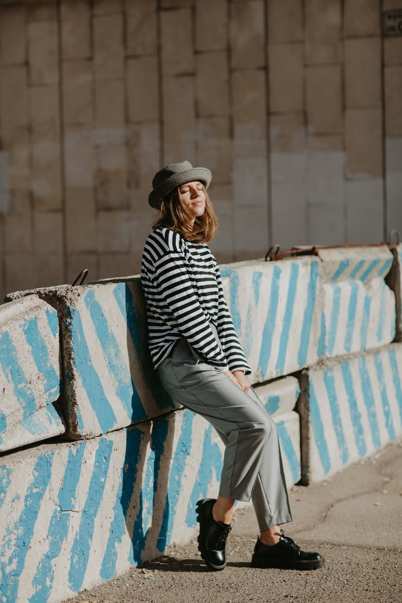 May include: A woman wearing a gray hat, black and white striped long-sleeve shirt, gray pants, and black shoes is standing in front of a concrete barrier with blue stripes.