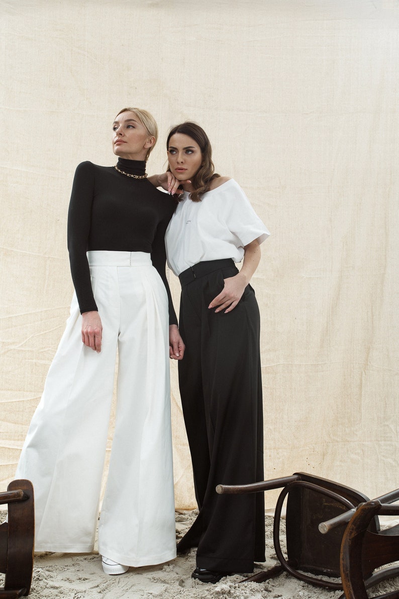 May include: Two women wearing wide-leg pants, one in white and one in black, stand in front of a beige backdrop. The woman in white is wearing a black turtleneck sweater and a gold chain necklace. The woman in black is wearing a white t-shirt. Both women are looking to the left of the frame.