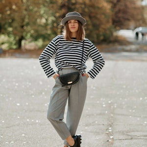 May include: A woman wearing a black and white striped long-sleeve shirt, gray pants, a gray fedora, and black shoes. She is carrying a black fanny pack.