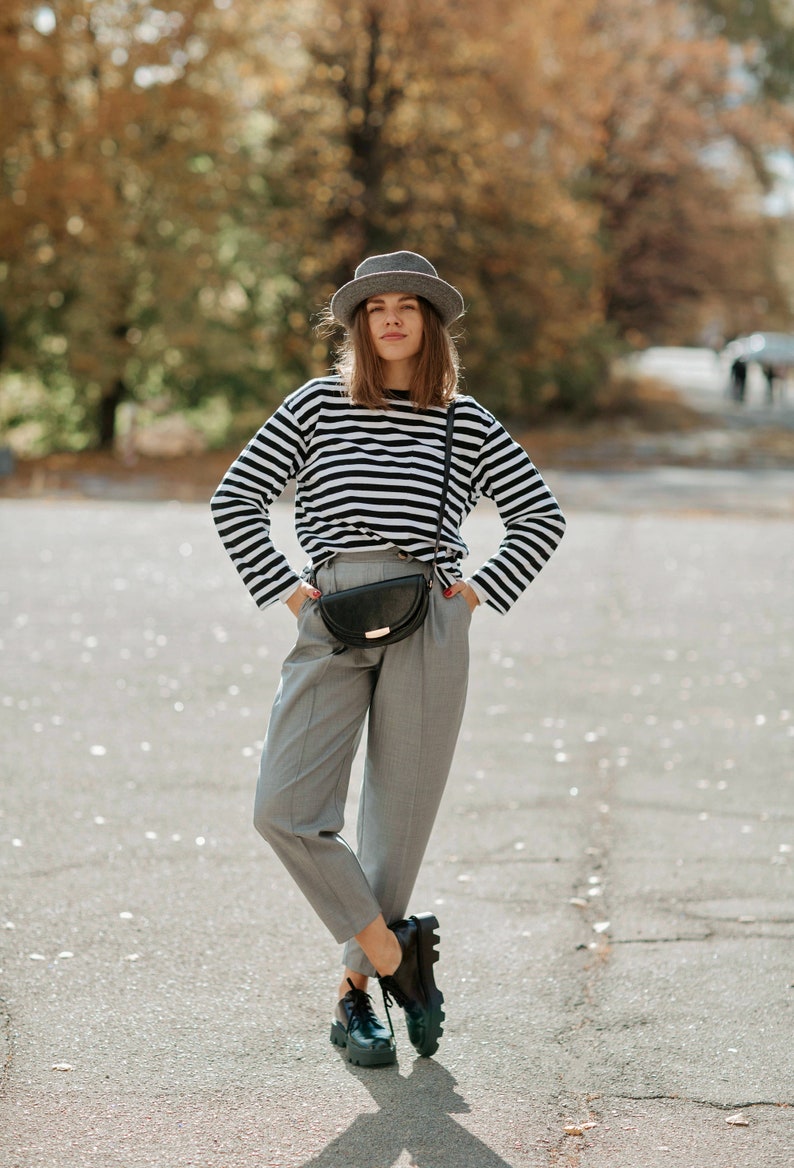 May include: A woman wearing a black and white striped long-sleeve shirt, gray pants, a gray fedora, and black shoes. She is carrying a black fanny pack.