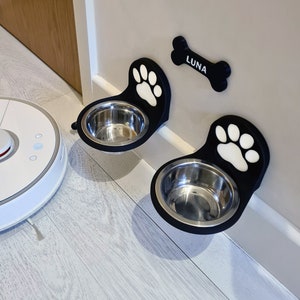 May include: Two black pet food bowls with paw print designs are mounted on a wall. The bowls are made of stainless steel and are attached to the wall with a black plastic bracket. The name "LUNA" is written on a bone-shaped plaque above the bowls.