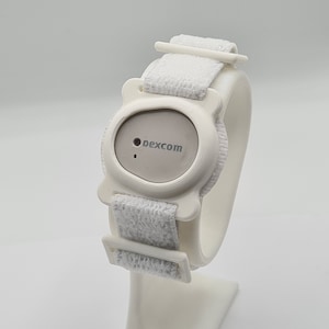 May include: A white medical device with a grey centre, featuring the text "dexcom." The device has a white strap with a hook-and-loop closure. The device is designed to be worn on the wrist.