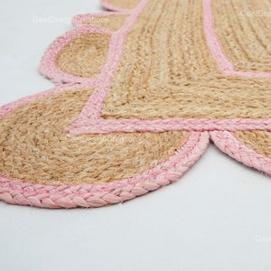 May include: A pink and beige jute rug with a scalloped edge. The rug is made of natural fibers and has a rustic, handmade look.