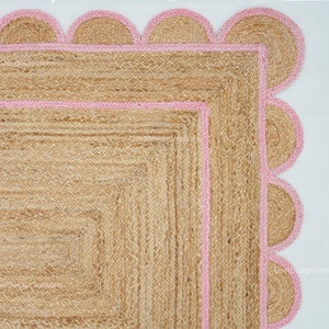 May include: A square jute rug with a pink scalloped border. The rug has a woven texture and is made of natural fibres.