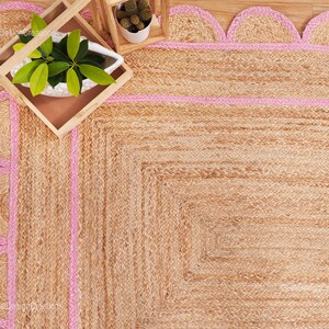May include: A natural fiber rug with a pink border. The rug has a scalloped edge and is made of jute.