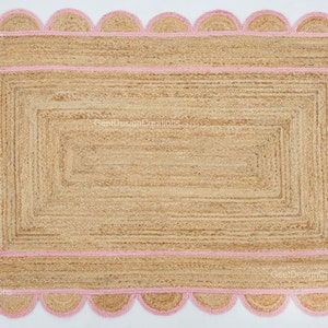 May include: A natural fiber rug with a pink scalloped border and a woven design. The rug has a rectangular shape and is made of jute.