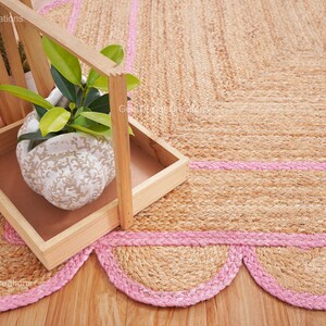 May include: A natural fiber rug with a pink border and scalloped edges. The rug is made of jute and has a natural, earthy color. A small potted plant sits on a wooden tray in the corner of the rug.