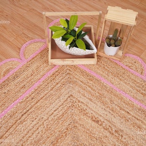 May include: A natural fiber rug with a pink border and a scalloped edge. The rug is made of jute and has a chevron pattern. Two potted plants are in the background.