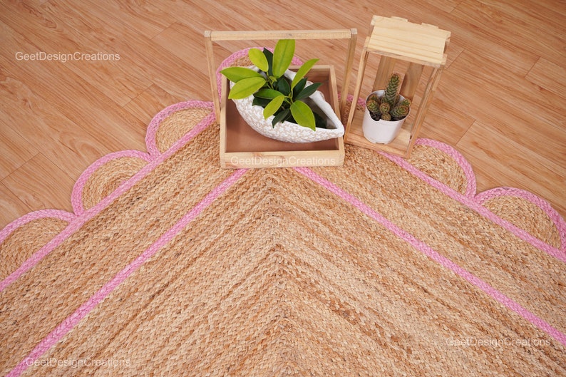 May include: A natural fiber rug with a pink border and a scalloped edge. The rug is made of jute and has a chevron pattern. Two potted plants are in the background.