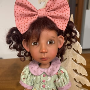 May include: A handmade doll with brown curly hair and a large pink bow with brown polka dots. The doll wears a green dress with a pink collar and cuffs. The doll has a serious expression.
