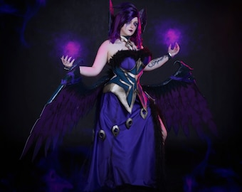 May include: A cosplayer in a dark purple and teal fantasy costume with large black feathered wings and purple hair. The costume includes a long purple dress, a teal and gold bodice, and is surrounded by purple smoke.