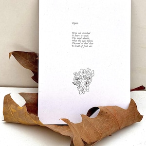 以下が含まれることがあります： 「Open」の文字と黒インクで印刷された詩が書かれた白いカード。花柄の装飾的な鍵穴のデザインが下部にあります。カードは乾燥した茶色の葉の上に置かれています。