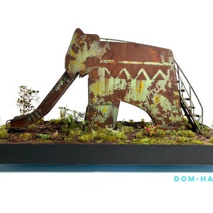 May include: A miniature, weathered playground slide in shades of rusty brown and green. The slide is set on a base with miniature plants and foliage. The text "DOM-HARDY" is visible at the bottom.