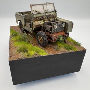 May include: A detailed model of a vintage, olive-green off-road vehicle with a weathered appearance. The model sits on a diorama base with grass and dirt, showcasing a realistic terrain. The vehicle's hood is open, revealing engine details.