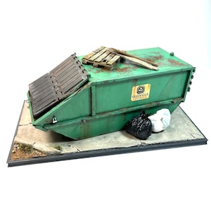 May include: A green recycling bin with a closed lid and a sign that says "Recycle Zero Waste". The bin is sitting on a grey concrete base with a black border. There are two black bin bags and a white plastic bag on the ground in front of the bin.