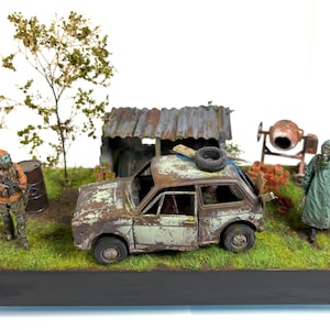 May include: A detailed miniature diorama of a post-apocalyptic environment. It includes a dilapidated car, figures in protective suits, a rusty shelter, and a tree. The scene is set on a grassy base with a black border.