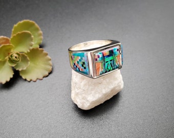 May include: A silver ring with a rectangular inlay of turquoise, coral, and other colorful stones. The ring is set on a white rock.