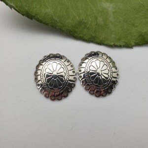 May include: Two silver-toned metal embellishments with a decorative floral design. Each embellishment has a scalloped edge and a raised center with a flower pattern.