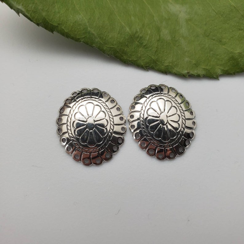 May include: Two silver-toned metal embellishments with a decorative floral design. Each embellishment has a scalloped edge and a raised center with a flower pattern.