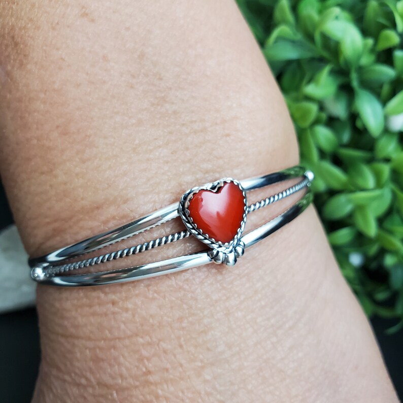 Op de afbeelding: Een zilveren armband met een rode hartvormige steen. De armband heeft drie banden, waarvan er twee gedraaid zijn.