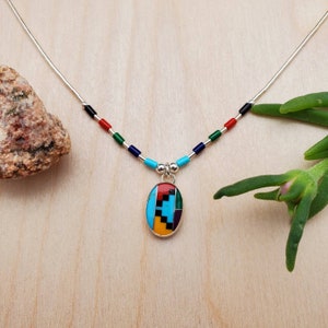 Puede incluir: Un collar de plata con un colgante ovalado multicolor. El colgante est&aacute; hecho de turquesa, rojo, amarillo, negro y verde. El collar est&aacute; enhebrado con peque&ntilde;as cuentas de color negro, rojo, verde y azul.