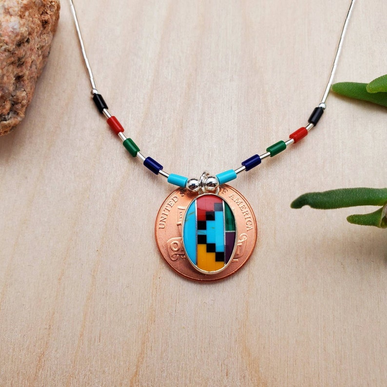 Puede incluir: Un collar de cadena de plata con cuentas de colores y un colgante con un dise&ntilde;o geom&eacute;trico hecho de piedras turquesa, rojo, amarillo, negro y verde. El colgante est&aacute; unido a una moneda de cobre con las palabras "United States of America" inscritas en ella.