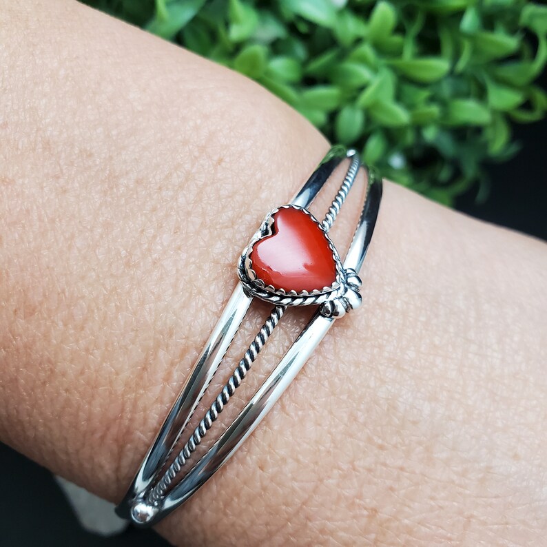 Op de afbeelding: Een zilveren armband met een rode hartvormige steen in het midden. De armband heeft een gedraaid draadontwerp.