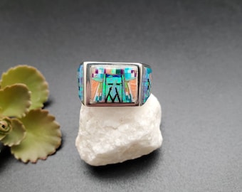 May include: A silver ring with a turquoise, pink, and green inlay design. The ring is set on a white rock.